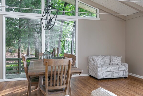 A dining area with a wooden table and chairs, a patterned sofa, and large windows offering a view of trees.