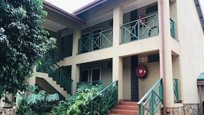 Un edificio con una escalera que conduce a un balcón, rodeado de follaje y un muro de piedra.