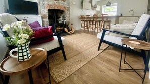 A living room with a stone fireplace, a TV, a sofa, and a dining area with bar stools. - Morfach Charming Modern Cottage within Conwy walls (Morfach)