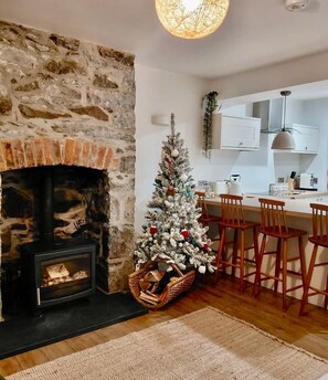 A cozy living room with a stone fireplace, a decorated Christmas tree, and a kitchen area with white cabinets and wooden chairs.