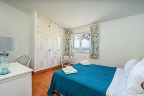 Quarto com cama, uma cadeira, escrivaninha e janela com cortinas.