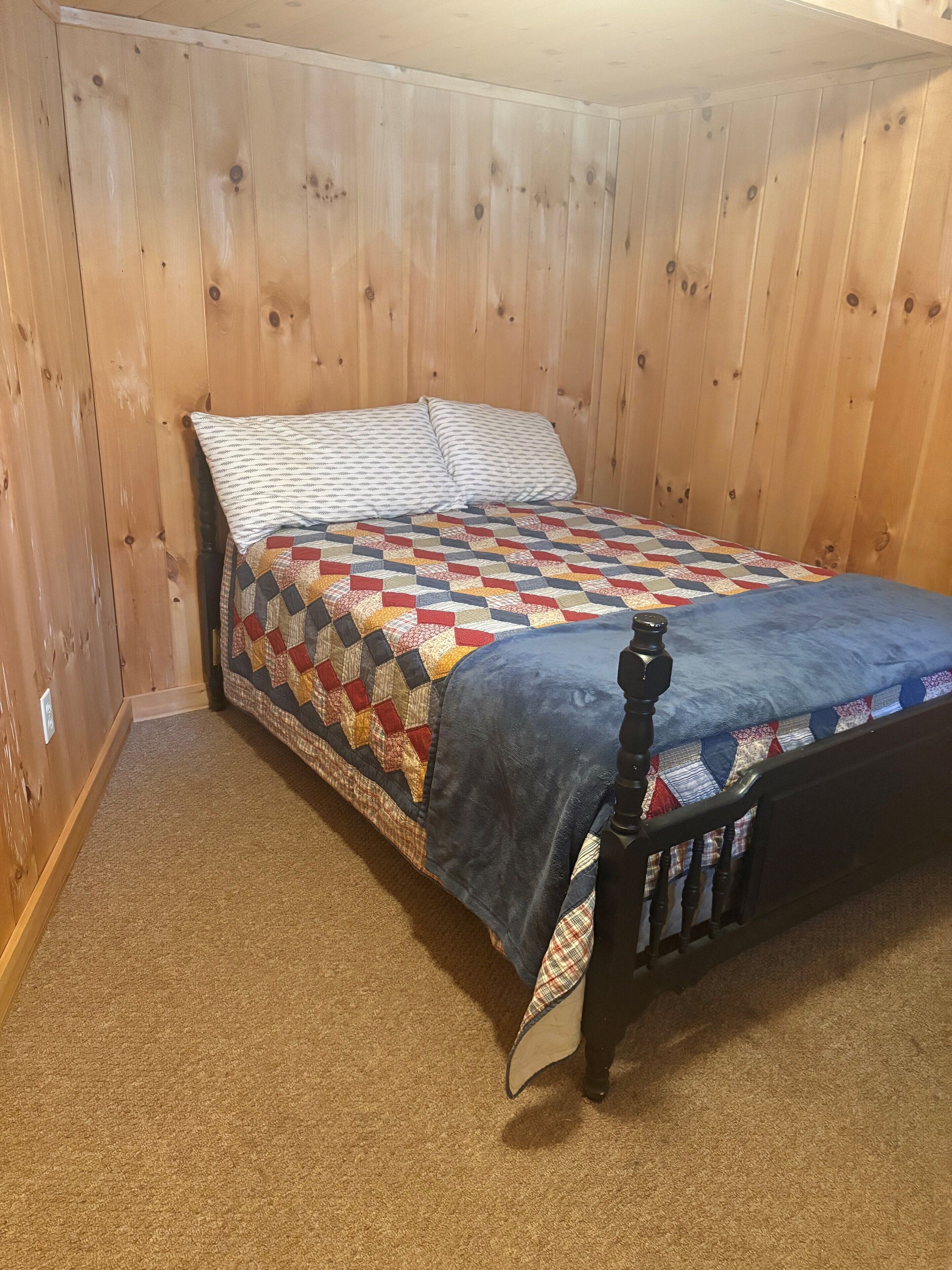 A single bed with a patterned quilt and pillows, a wooden headboard, and a carpeted floor in a room with wooden walls.