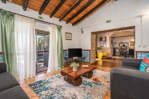 Ampla sala de estar com teto de madeira, televisão, mesa de centro com uma planta e vista para a área de jantar.
