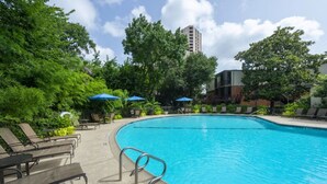 Piscina com espreguiçadeiras e guarda-sóis, rodeada por vegetação e um edifício alto ao fundo.