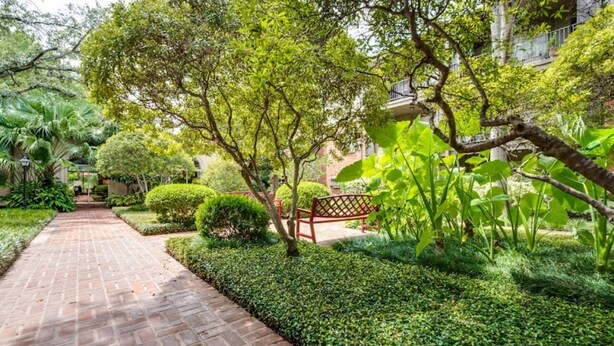 Uma trilha de tijolos serpenteia por um jardim exuberante, com sebes bem aparadas e árvores.