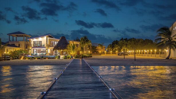 Una propietat a primera línia de mar amb un camí de fusta que condueix a una zona de piscina, envoltada de palmeres i il·luminada amb llums ambientals.