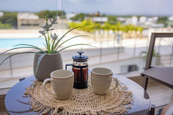 A table with a coffee maker, two mugs, and a potted plant on a woven placemat, overlooking a pool and greenery.