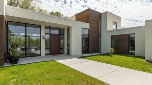 A modern building with large glass doors, a wooden facade, and a well-maintained lawn. - Modern, Ultra-Luxurious, 5 BR/4.5 BA Home on a quiet wooded lake in Ann Arbor (Superior Township)