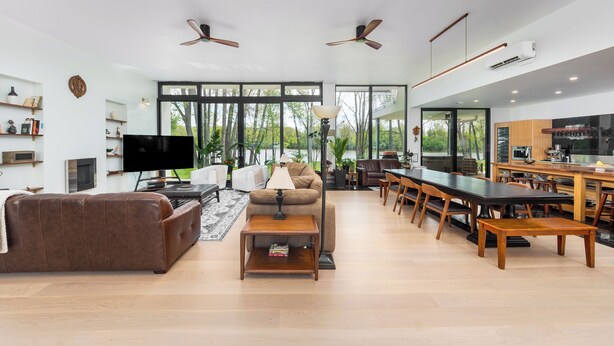 A spacious living room with a brown leather sofa, a wooden coffee table, and a TV. There are ceiling fans, a kitchen area with a dining table, and large windows offering a view of greenery outside. - Modern, Ultra-Luxurious, 5 BR/4.5 BA Home on a quiet wooded lake in Ann Arbor (Superior Township)