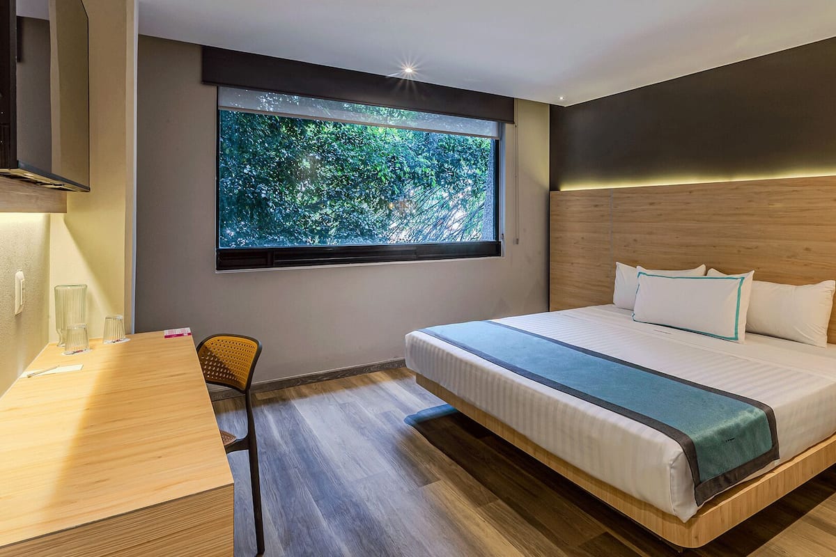 A modern hotel room with a bed, a desk, a chair, and a large window with a view of trees.