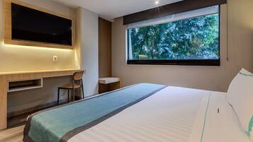 A hotel room with a bed, a desk, a chair, a TV, and a window with a view of trees.