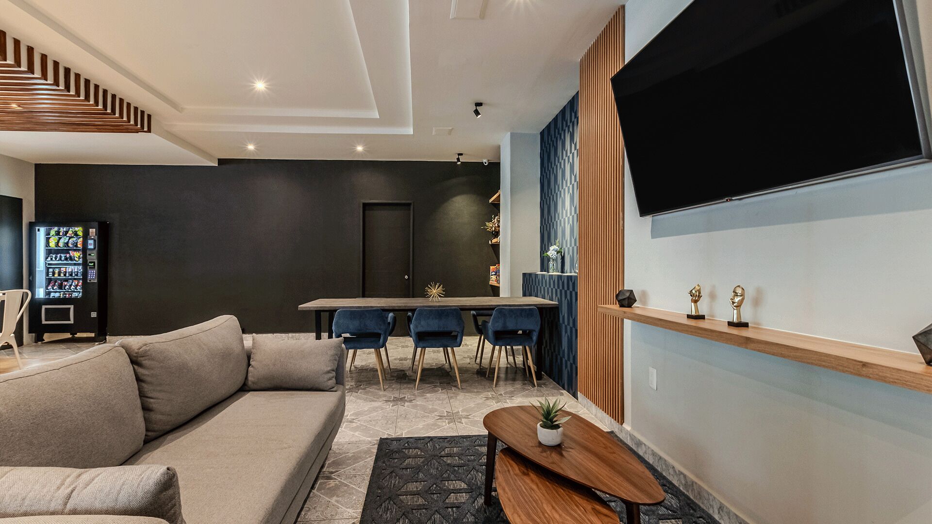 A modern living room with a sofa, a dining table with chairs, a TV mounted on the wall, and a vending machine.