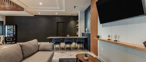 Una sala de estar moderna con un sofá, una mesa de comedor con sillas, un televisor montado en la pared y una máquina expendedora.