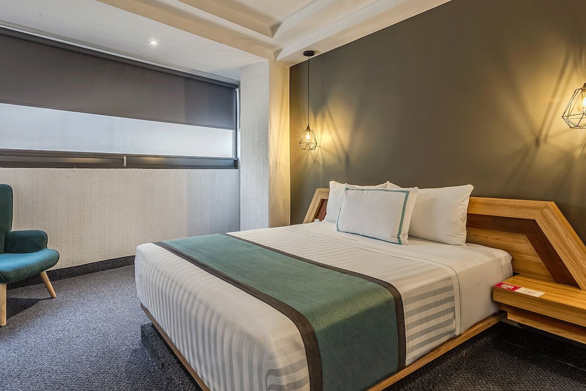 A hotel room with a bed, a chair, a wall-mounted light, and a window.