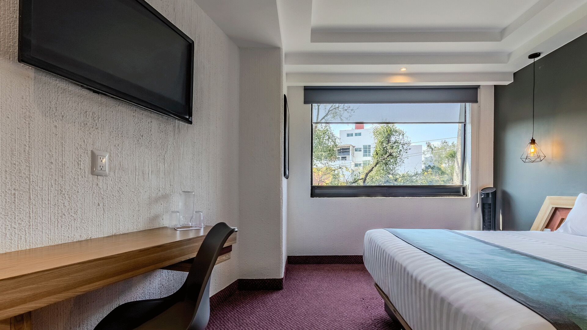 A bedroom with a bed, a desk, a chair, a TV mounted on the wall, and a window with a view of buildings and trees.