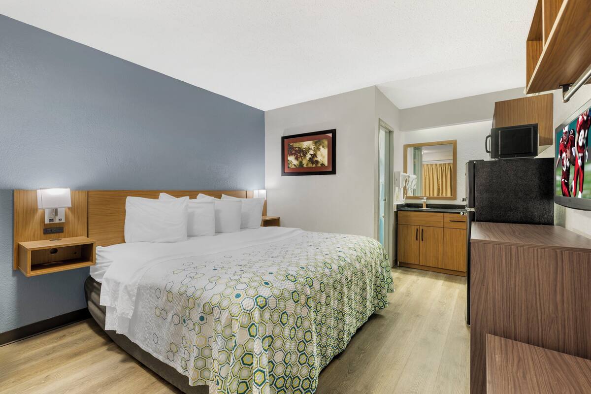 A hotel room with a large bed, a wooden headboard, a nightstand with a lamp, a small wooden cabinet, a microwave, and a wooden counter with a sink.