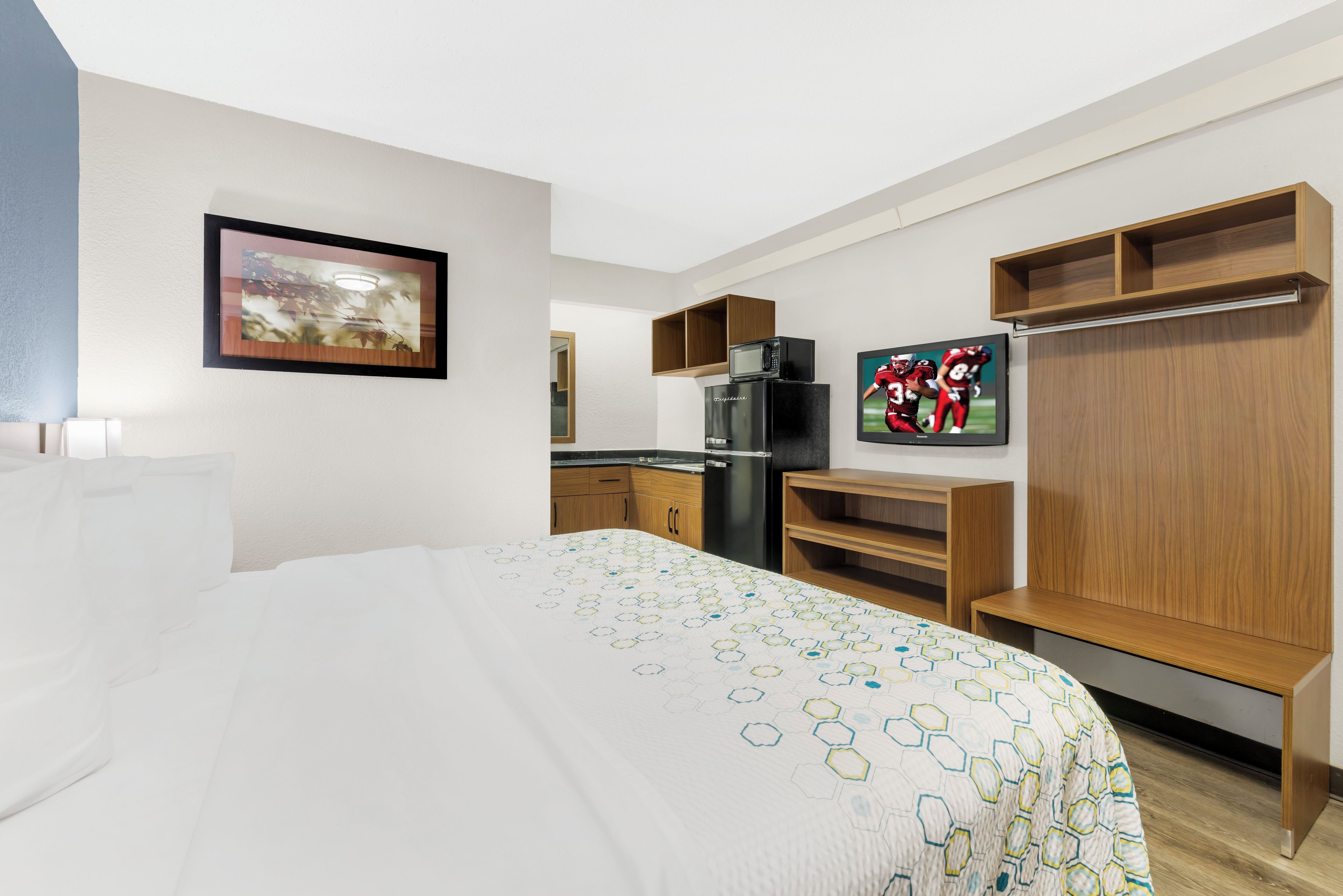 Habitación de hotel con cama, microondas, refrigerador, estantes de madera y un cuadro en la pared.