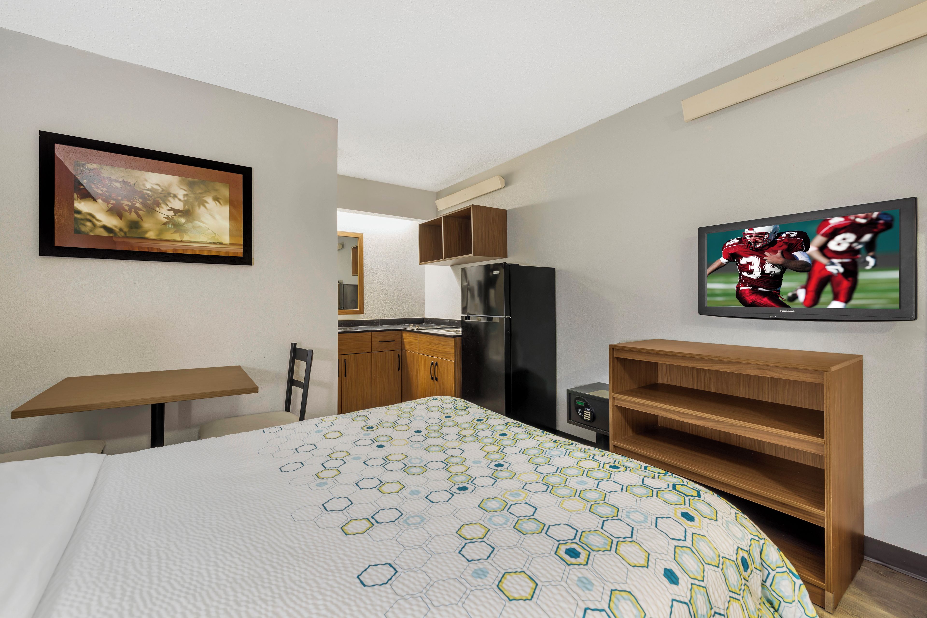Una habitación de hotel con cama, escritorio, refrigerador, televisor y un cuadro en la pared.