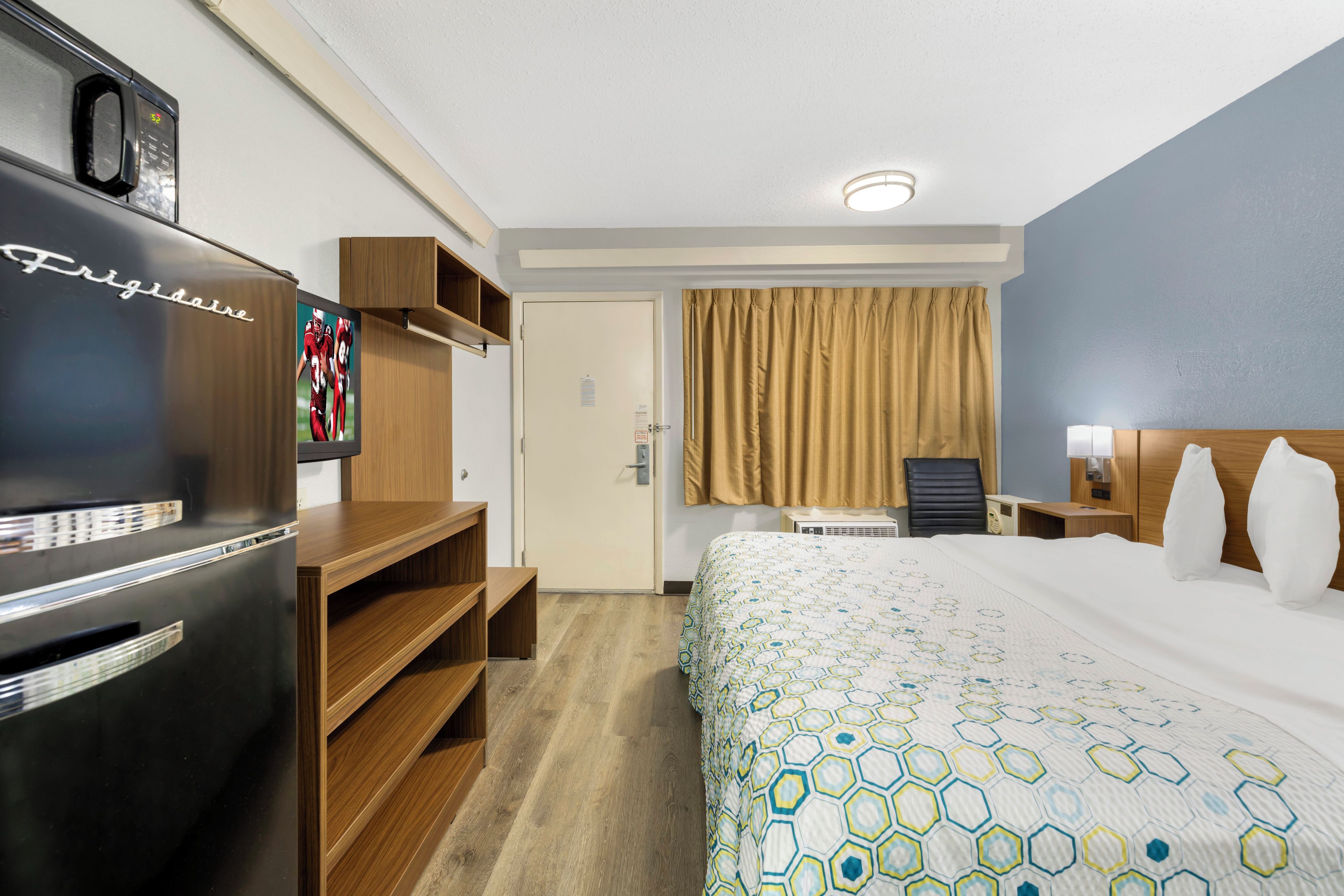 A hotel room with a bed, a desk, a refrigerator, a television, and a window with curtains.