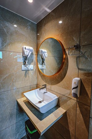 Un baño moderno con pared de mármol, un espejo redondo, un lavabo blanco y dos secadores de pelo montados en la pared.