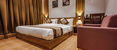 A hotel room with a large bed, wooden headboard, two bedside tables with lamps, a wooden nightstand, a red armchair, and a wooden floor.