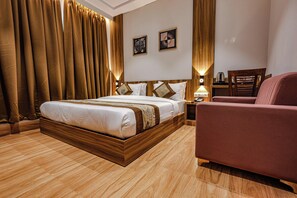 A hotel room with a large bed, wooden headboard, two bedside tables with lamps, a wooden nightstand, a red armchair, and a wooden floor.
