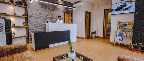 Zona de recepción de hotel con recepcionista, mostrador de recepción, una mesa auxiliar con planta y un reloj en la pared.