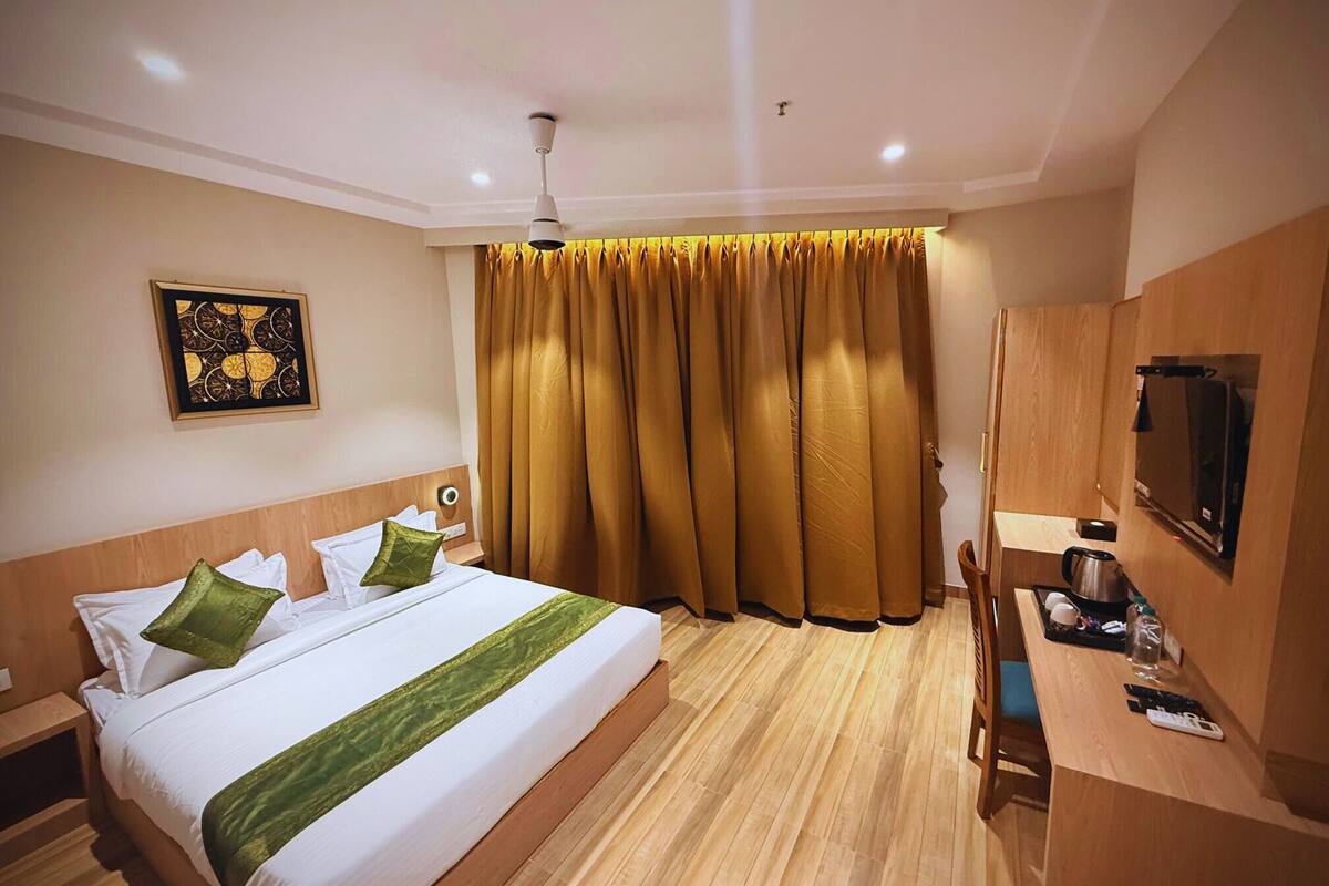 A hotel room with a bed, a desk with a kettle, a television, and a large window with curtains.