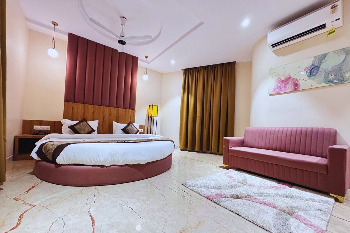 A bedroom with a round bed, a pink sofa, a ceiling fan, an air conditioner, and a large window with curtains.