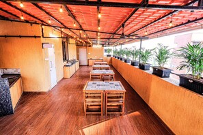 A covered outdoor seating area with wooden tables and chairs, potted plants along the wall, and a tiled floor.