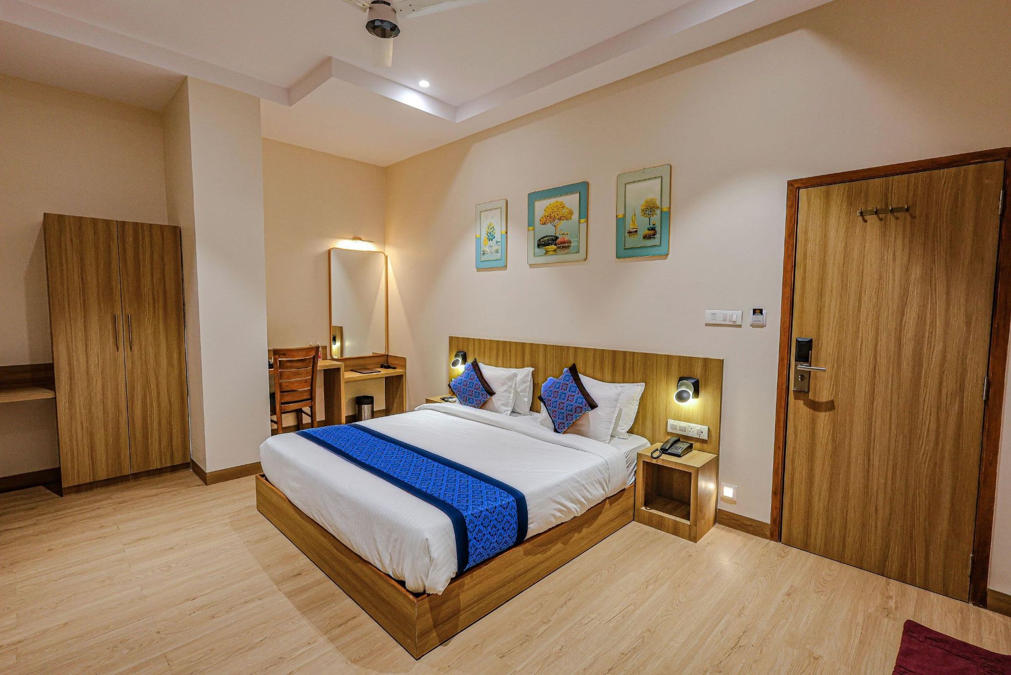 A hotel room with a wooden bed, a desk, a chair, a wardrobe, and three wall-mounted artworks.