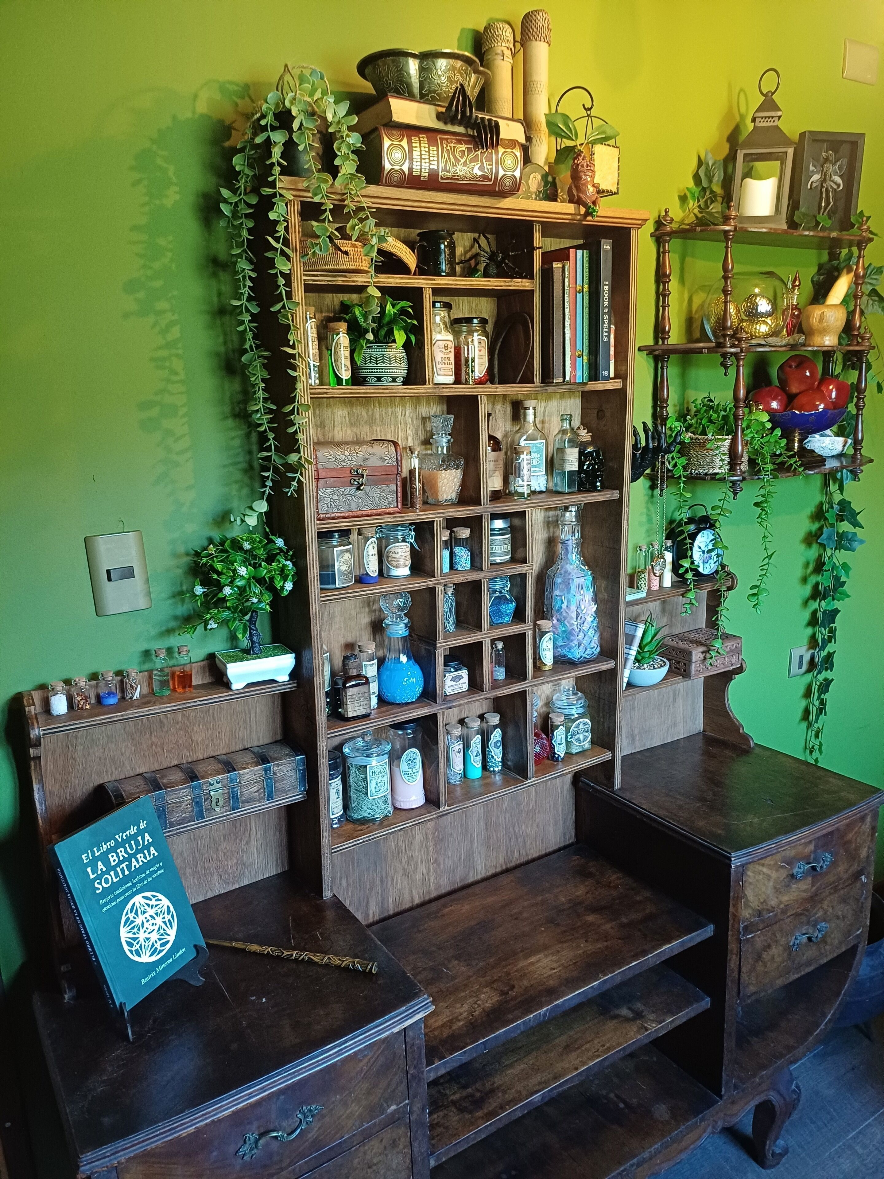 A room with a wooden bookshelf filled with various items, a small table with a book, and a green wall.