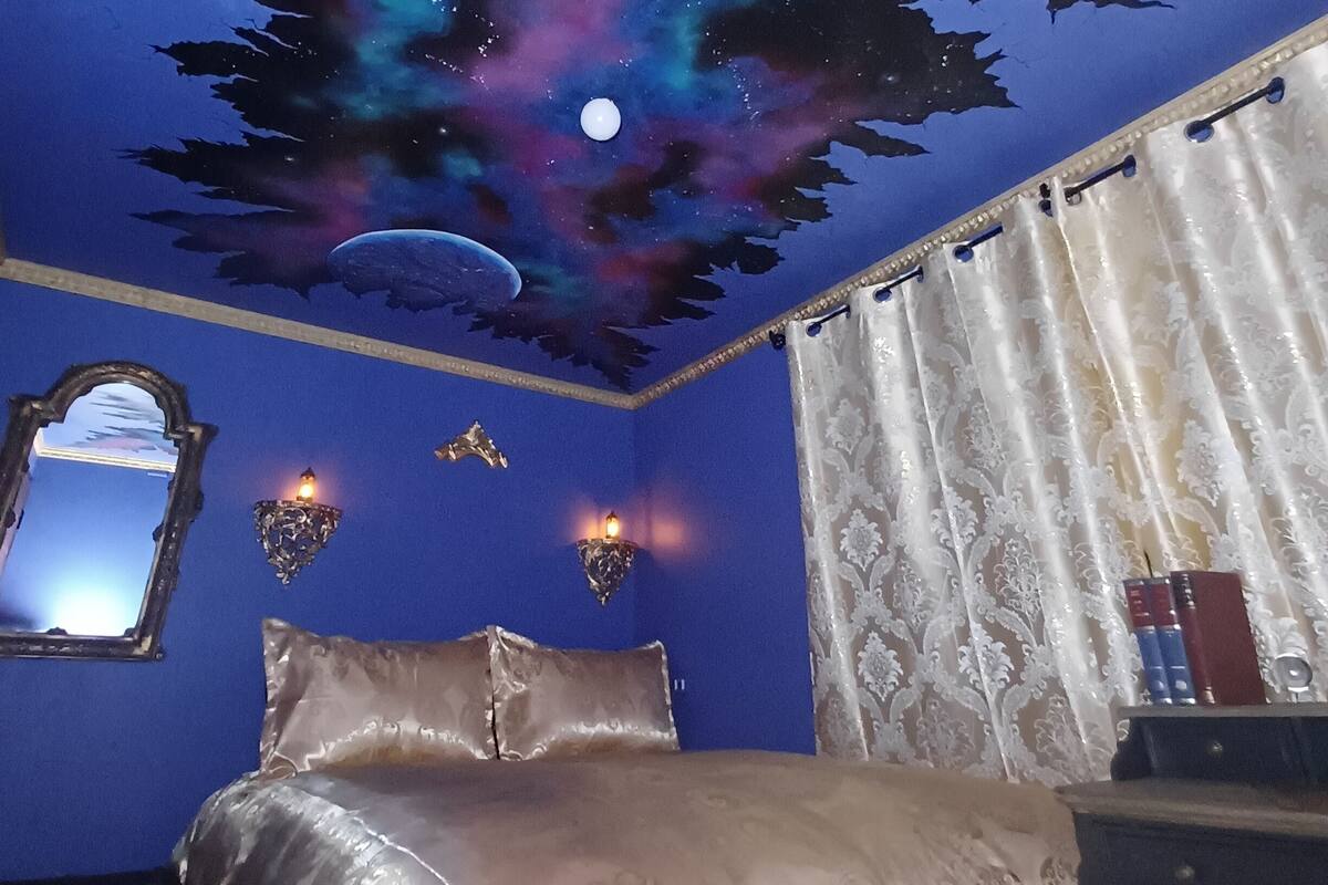 A bedroom with a bed, a night sky mural on the ceiling, a mirror, and a window with curtains.