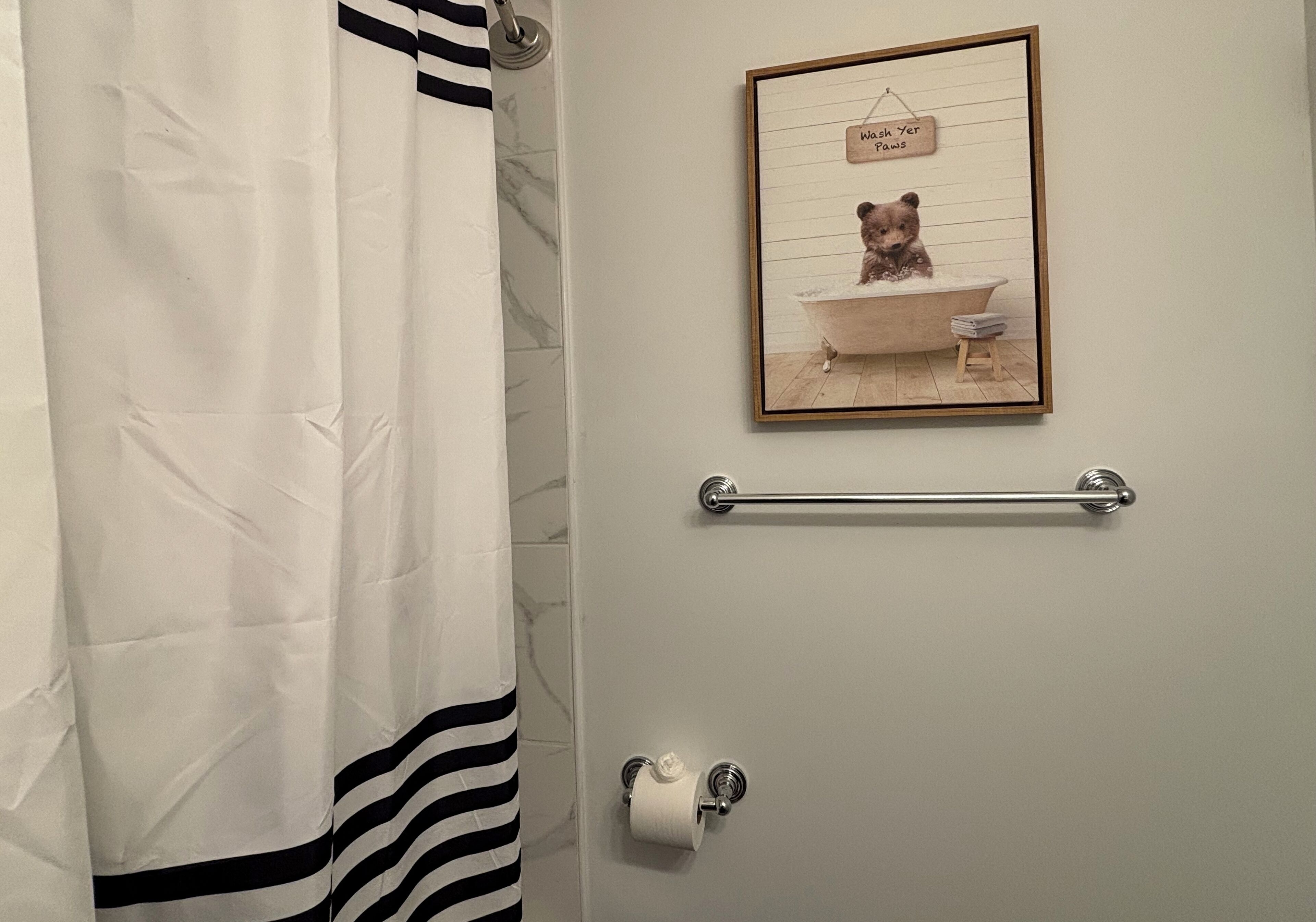 A bathroom with a striped shower curtain, a framed picture of a bear in a bathtub, a towel rack, and a toilet paper holder.