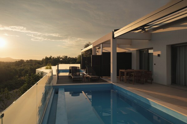 A modern outdoor pool area with lounge chairs and a dining table, surrounded by a white building with large windows. - VILA 2 ANAPNOE IKIES CORFU WITH PRIVATE POOL (Corfu)