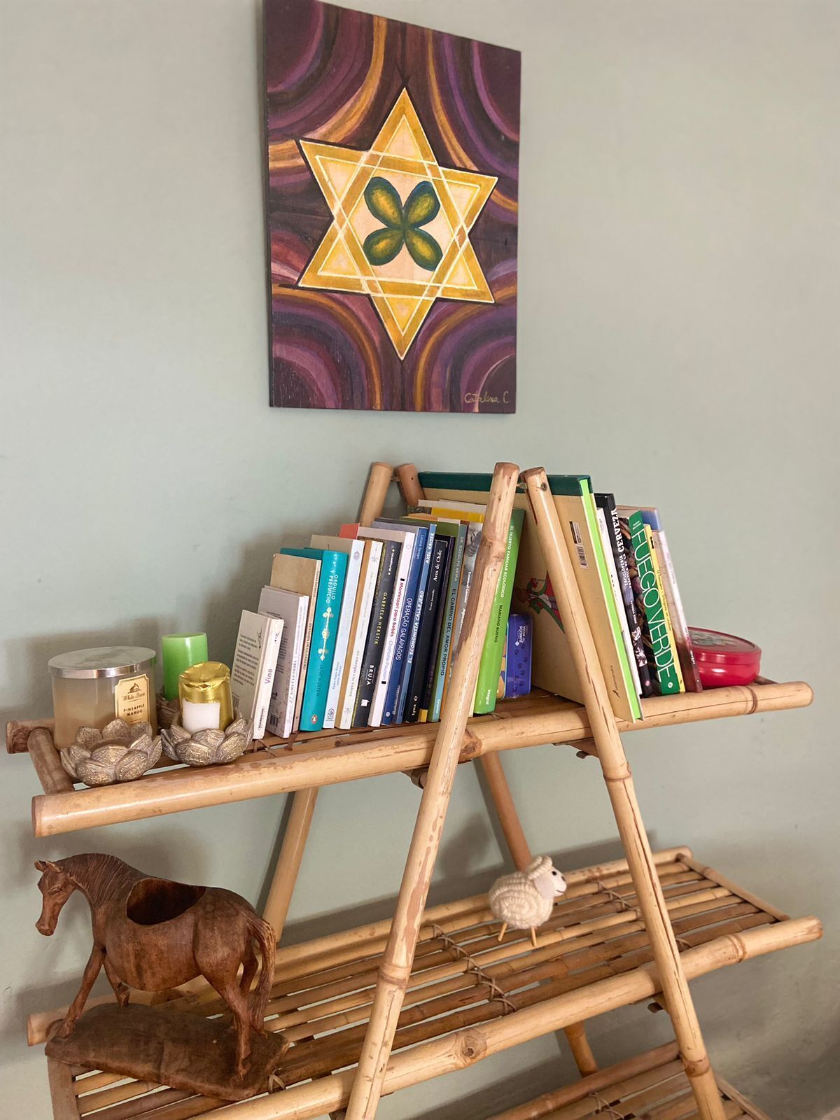 Una estantería de madera con libros y objetos decorativos, una escultura de caballo de madera y una pared con un cuadro de motivos geométricos.