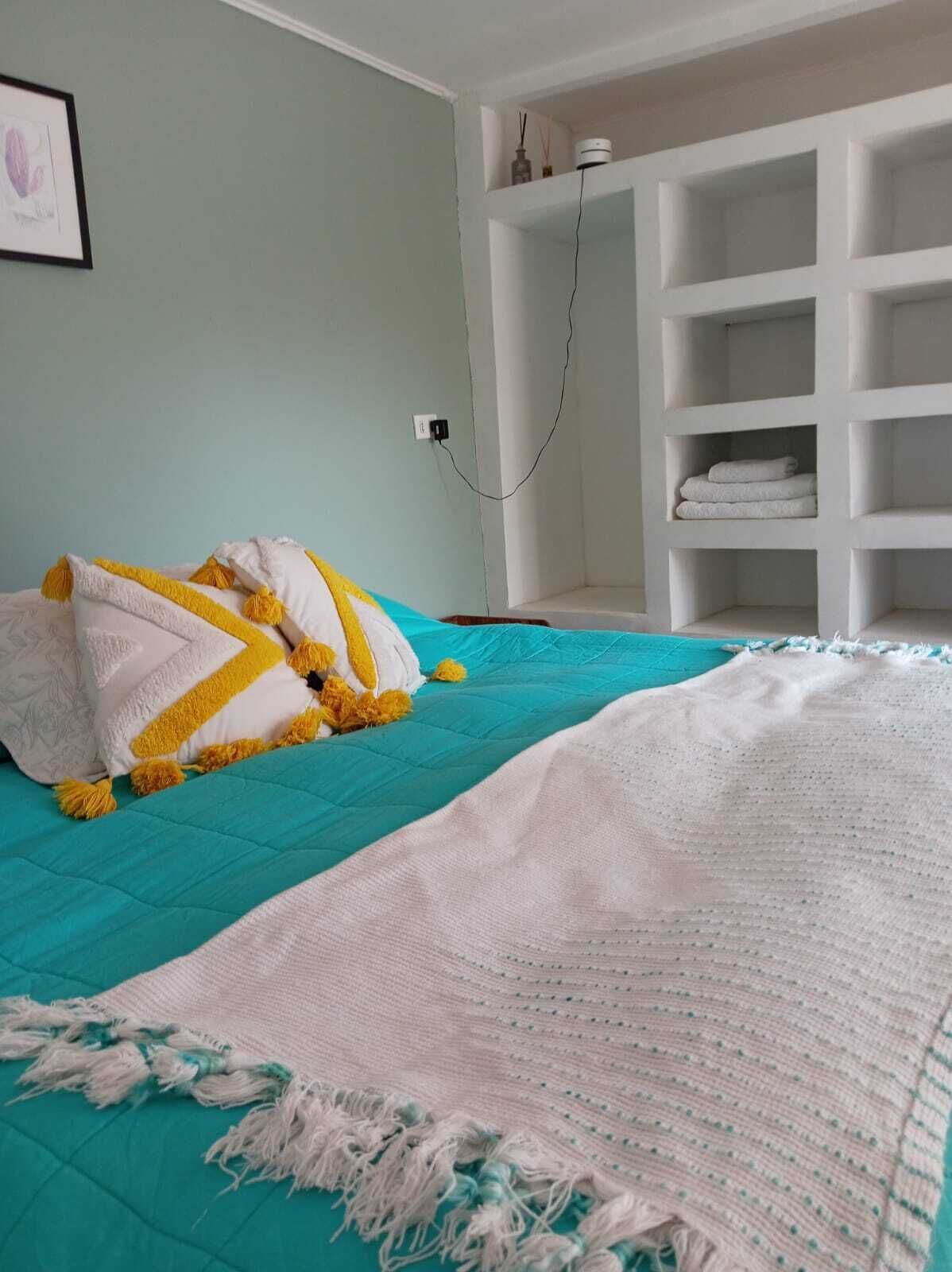 Une chambre avec une couvre-lit turquoise, des coussins blancs et jaunes, un plaid blanc en tricot et une étagère blanche adossée à un mur vert clair.