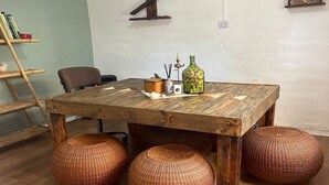 Une table à manger en bois, agrémentée de poufs tressés, une croix en bois fixée au mur et une petite étagère en bois sur laquelle est posée une photo.