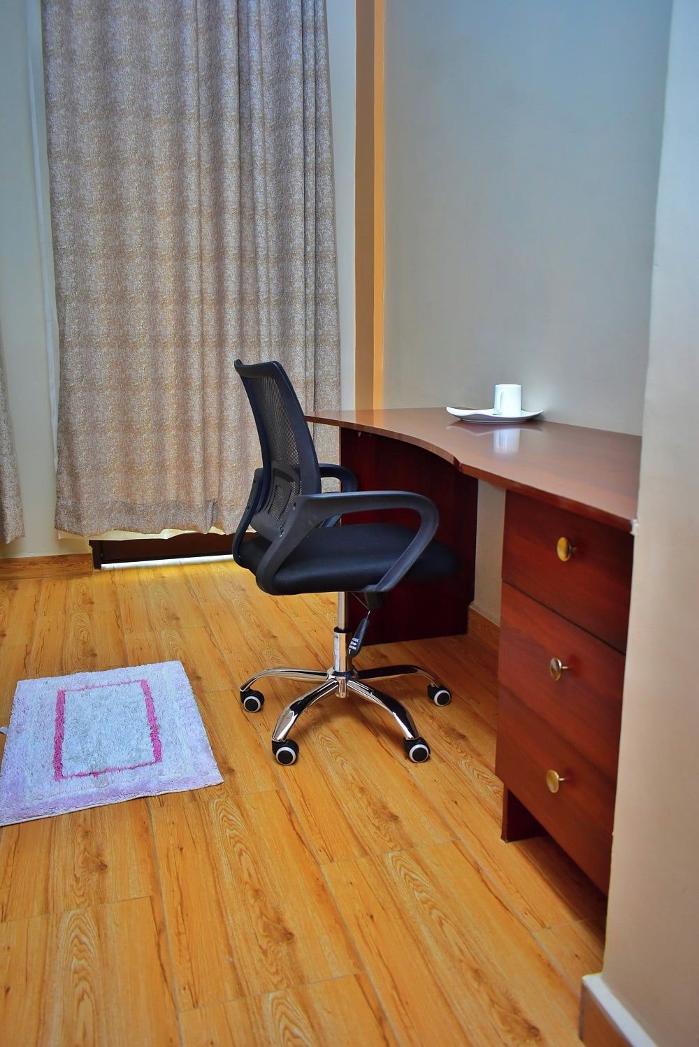 Une pièce avec un plancher en bois, un fauteuil de bureau noir, un bureau en bois doté d’un tiroir et un rideau.