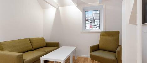 A modern, minimalistic living room with a white sofa, a matching armchair, and a white table. A window with a view of buildings is visible.