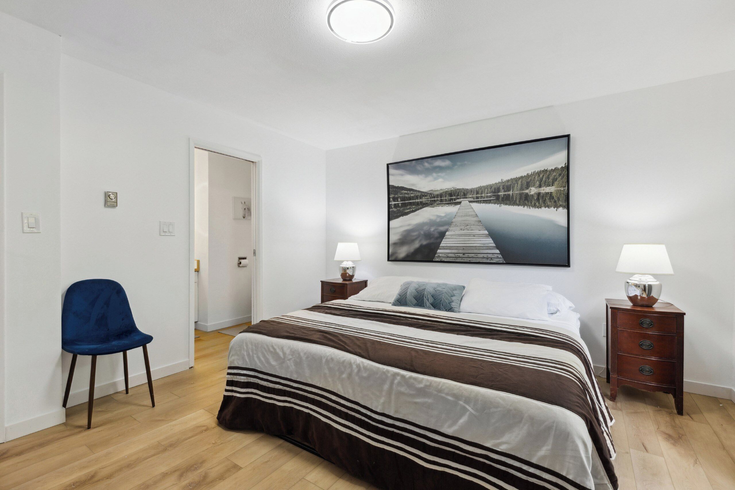 A bedroom with a large bed, a chair, a nightstand, and a wall-mounted picture.