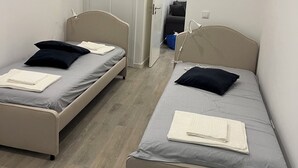 A small, modern room with two single beds, each with a pillow and a blanket. The room has a wooden floor and white walls.