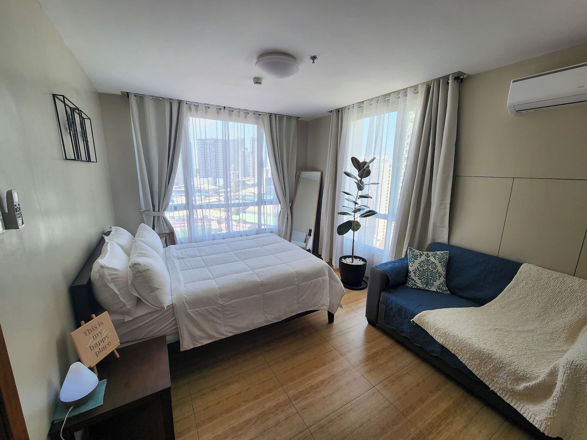 A modern hotel room with a bed, a sofa, a potted plant, and a view of the cityscape.