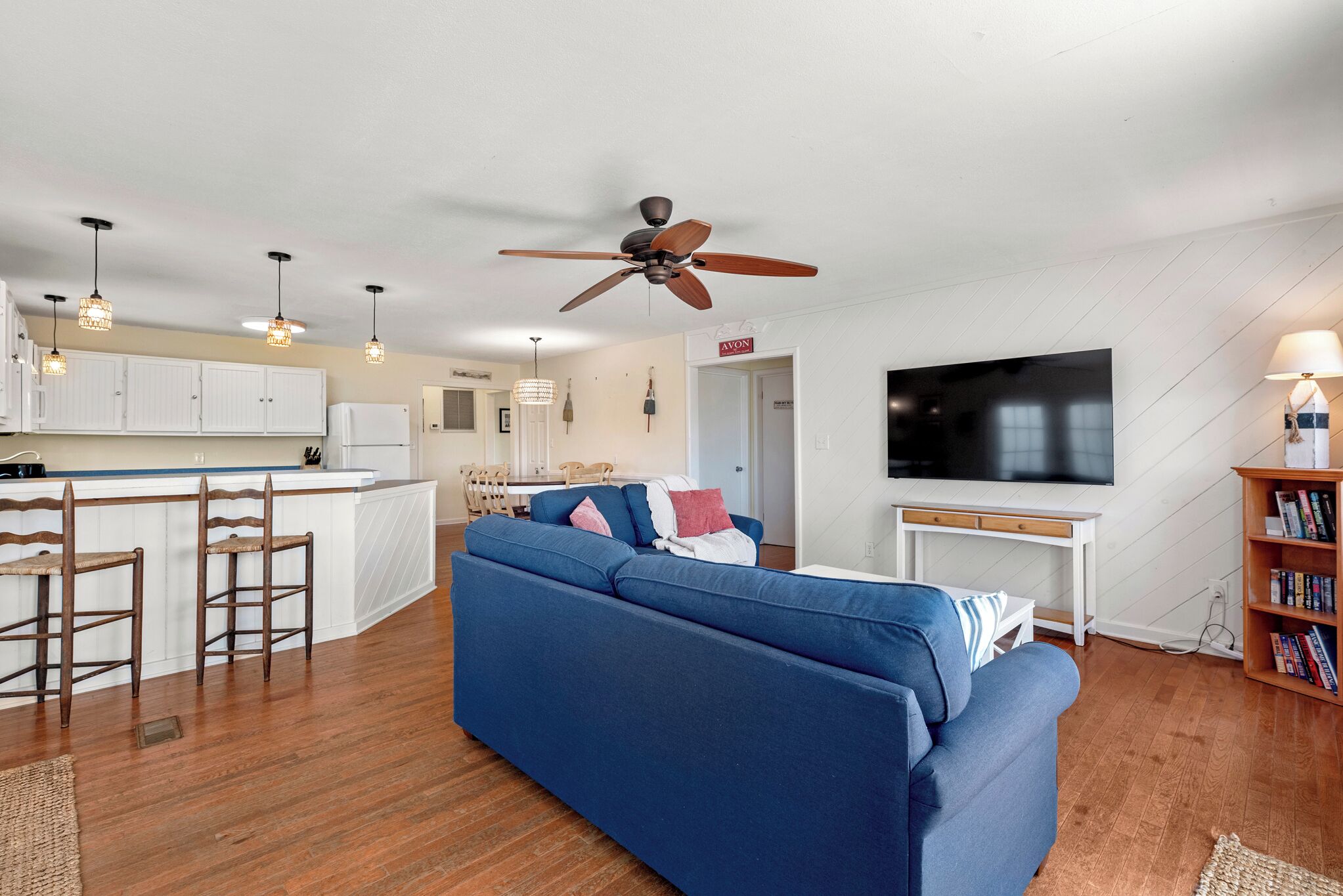 A modern living room with a blue sofa, wooden flooring, a ceiling fan, pendant lights, a TV, and a bookshelf.