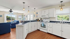 A modern kitchen with white cabinets, a built-in oven, and a large island. There is a ceiling fan and pendant lights above the island. - Surf'n Sound: Canalfront | Gas Grill (Avon)