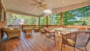 A covered outdoor deck with wicker furniture and a glass table, surrounded by wooden railings and trees. - Golf View Retreat Near Ski & Hiking Trails (Beech Mountain)