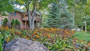 A brick house with a balcony, surrounded by a garden with orange flowers and a large rock in the foreground. - Golf View Retreat Near Ski & Hiking Trails (Beech Mountain)