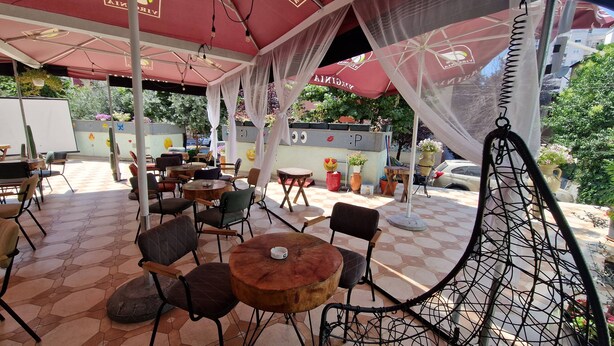 An outdoor cafe with round tables and chairs, covered by red umbrellas and white canopies, with potted plants and a decorative wall in the background. - Frekel Hotel (Tirana)