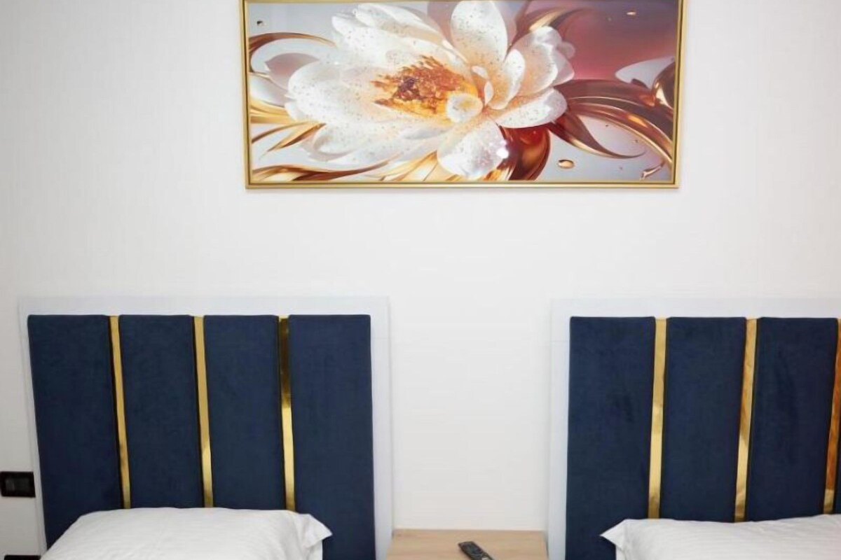A hotel room with two beds, each with a blue headboard and white bedding. There is a wooden nightstand between the beds with a remote control on it. Above the beds, there is a framed floral artwork on the wall.