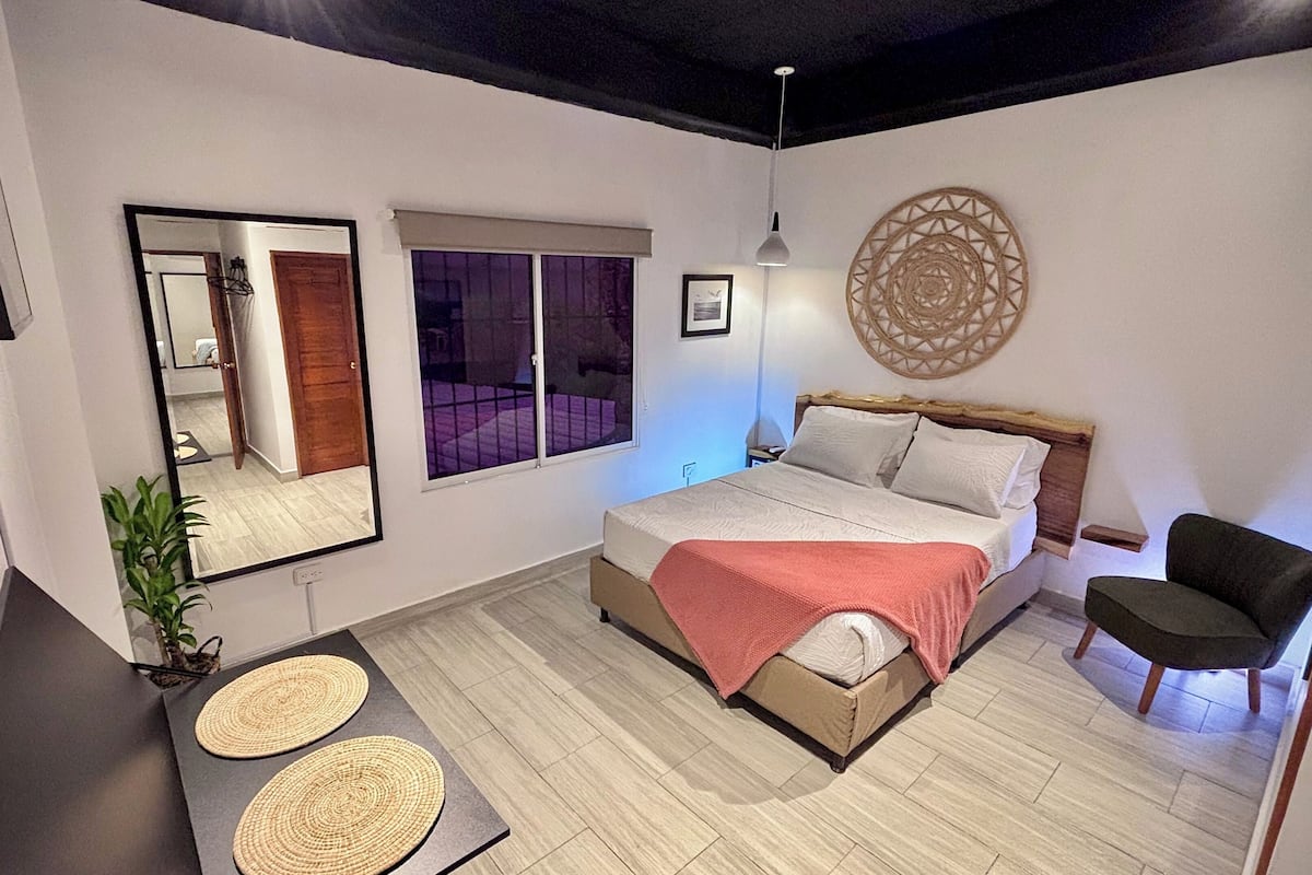 A bedroom with a bed, bedside table, chair, mirror, window, and wall decor.